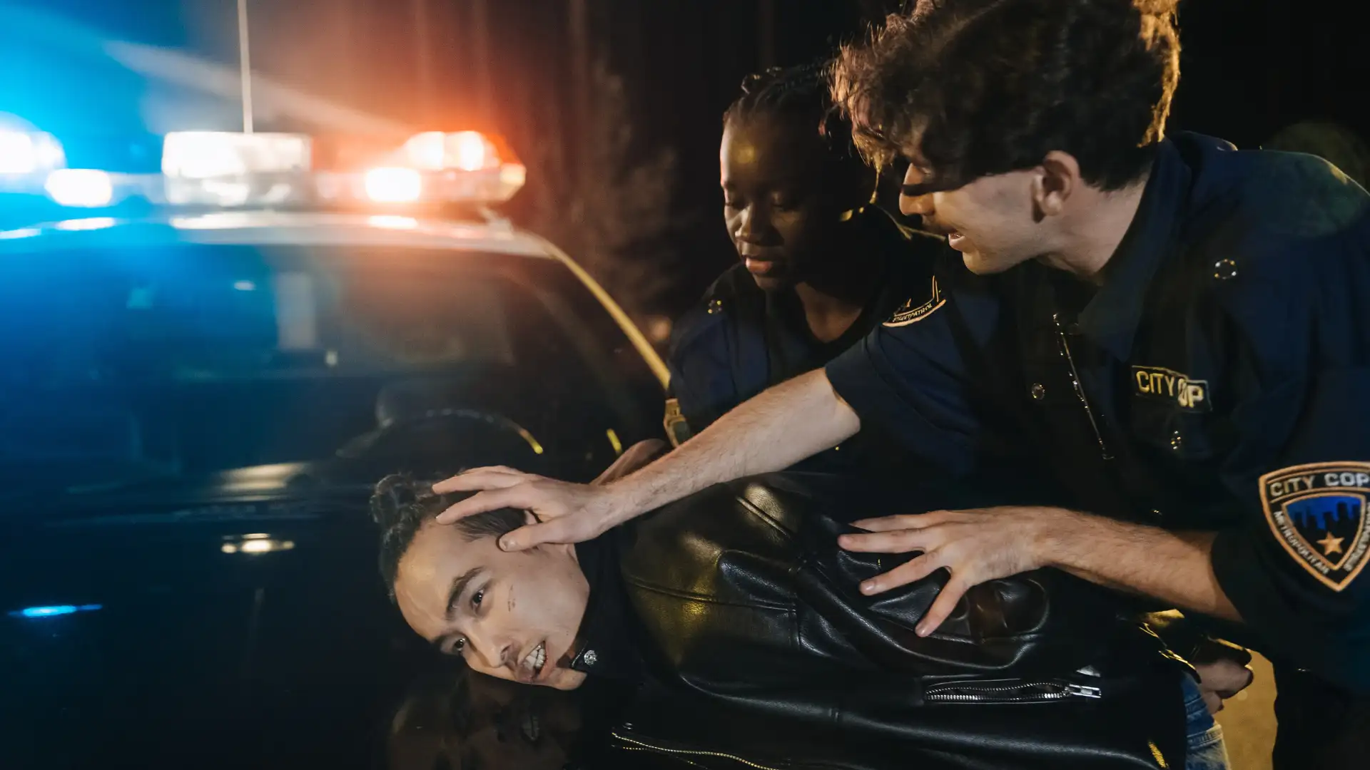 Police officers detain a person against a car at night with flashing lights.