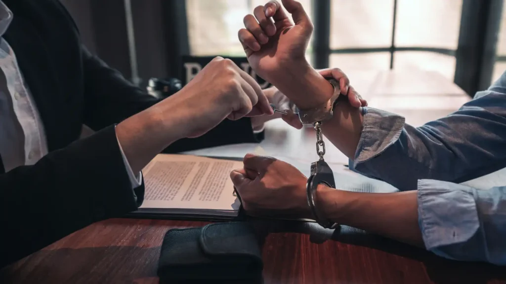 A person in handcuffs sitting across from another individual, possibly a lawyer, in a legal setting. 