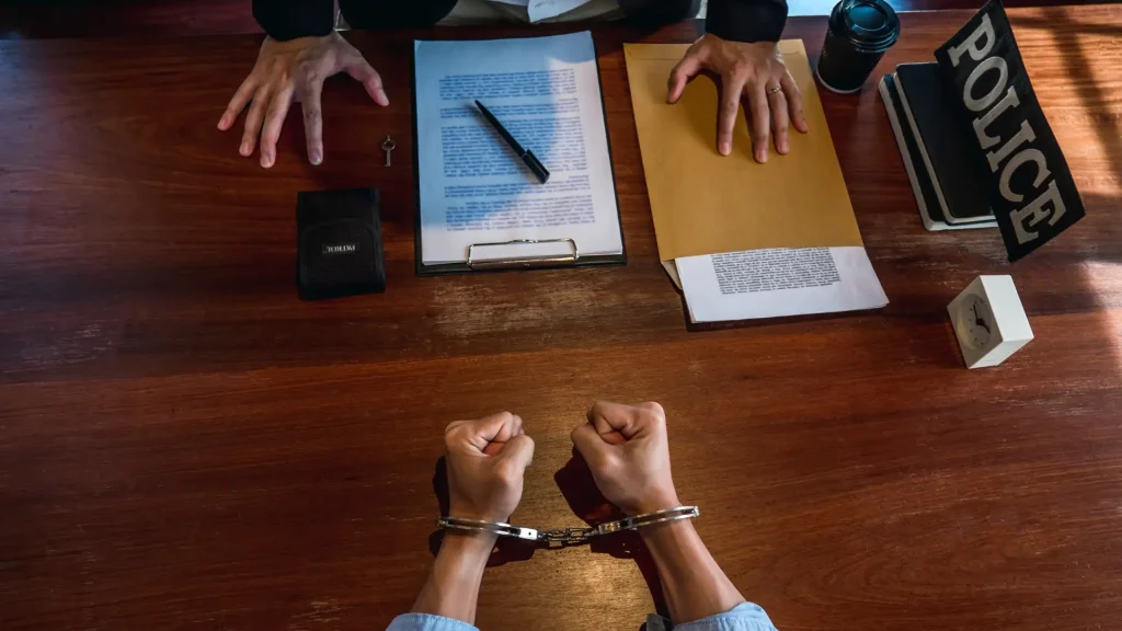A handcuffed person sits across from an officer at a desk with legal documents.