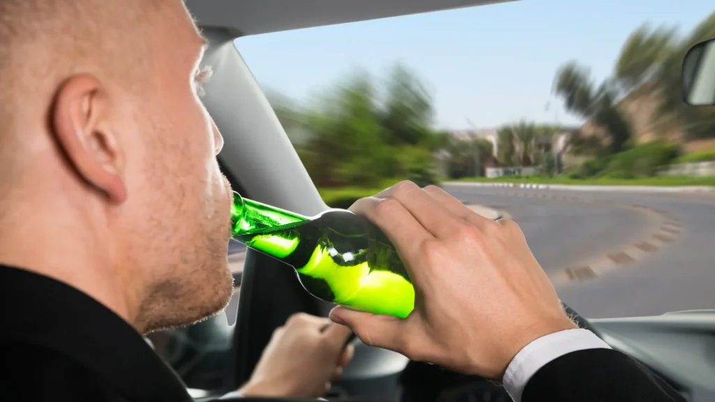 A person holding a green bottle while driving a car, indicating potential alcohol consumption behind the wheel.